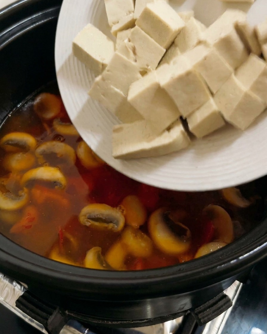Würfel aus festem weißem Tofu werden in eine köchelnde Suppe auf Tomatenbasis mit Pilzen gegeben.