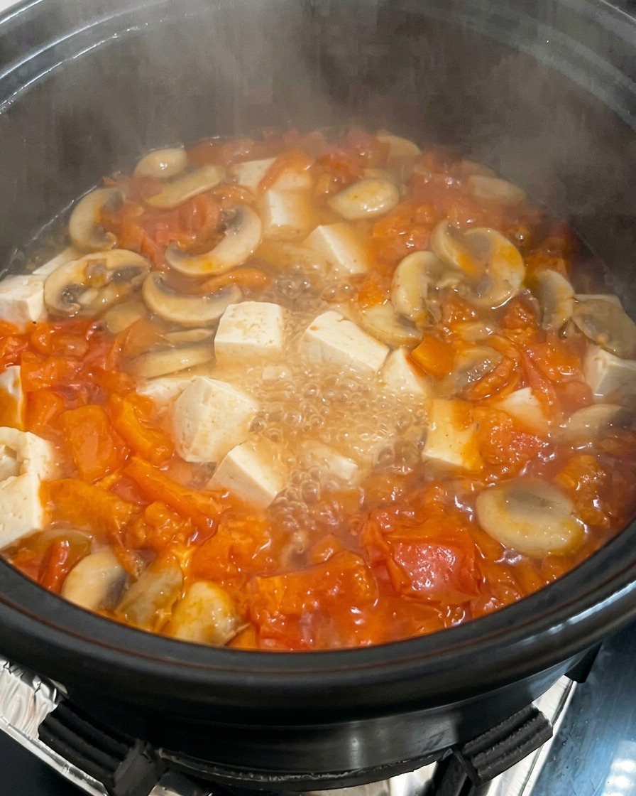 Ein Topf mit köchelnder Tomaten-Pilz-Suppe mit Tofu-Würfeln, in den ein verquirltes Ei geträufelt wird.