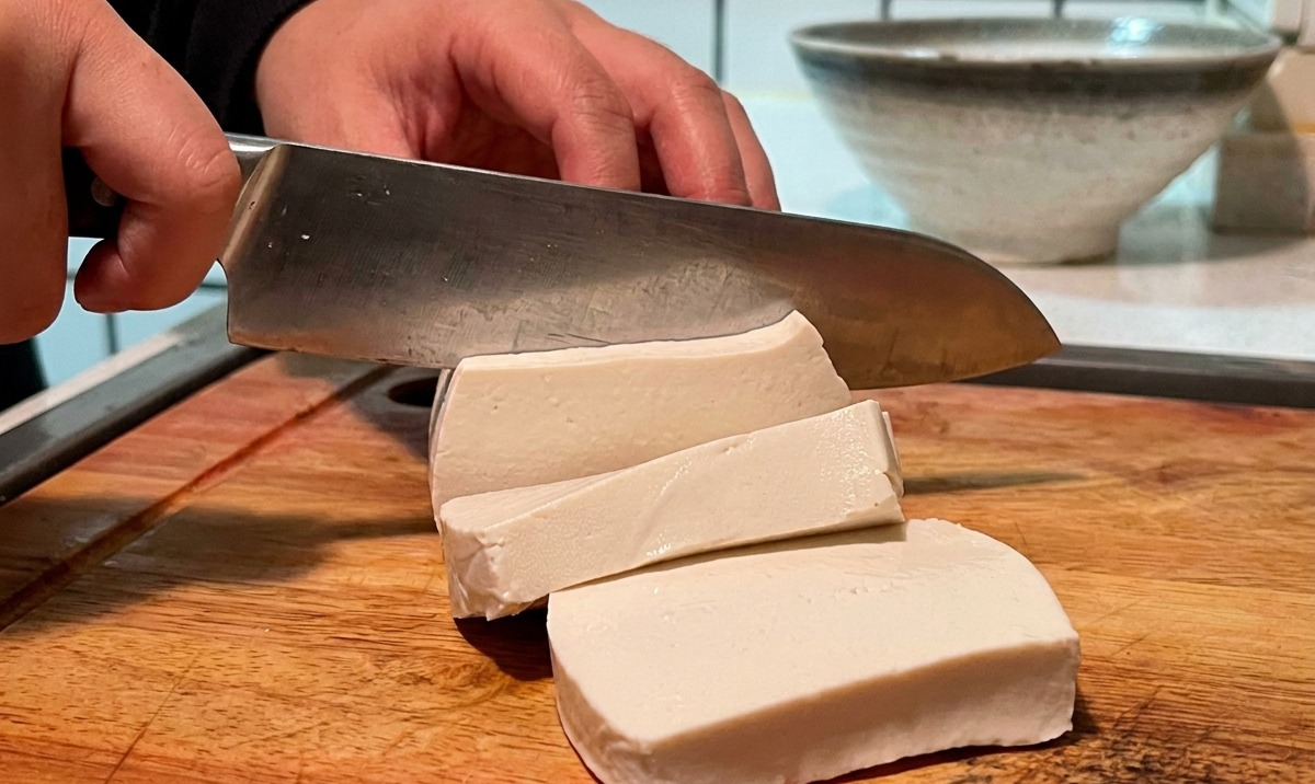 Ein Block weißer Tofu, der auf einem Holzbrett in kleine Würfel geschnitten wird.