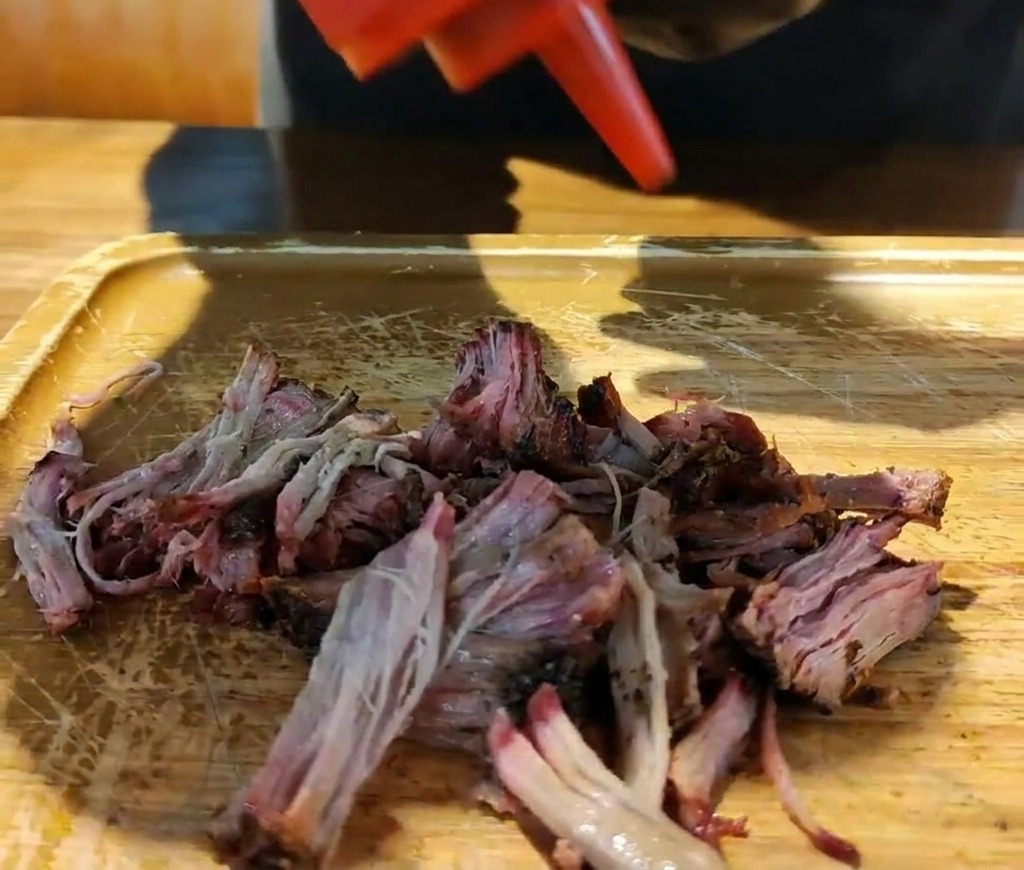 Eine rote Spritzflasche gießt dicke Barbecue-Sauce über einen Haufen zerzupftes, geräuchertes Schweinefleisch auf einem Holzbrett.