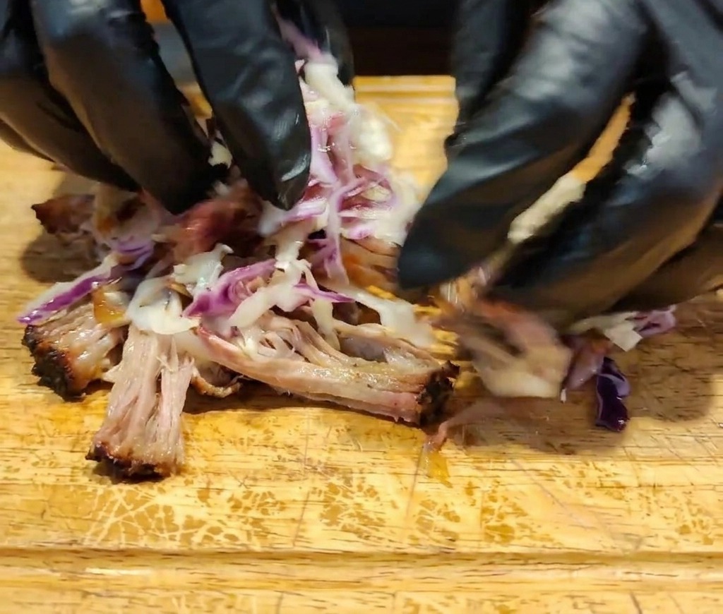 Hände vermischen das Fleisch, die dunkle Sauce und den bunten Coleslaw direkt auf einer Holzoberfläche.