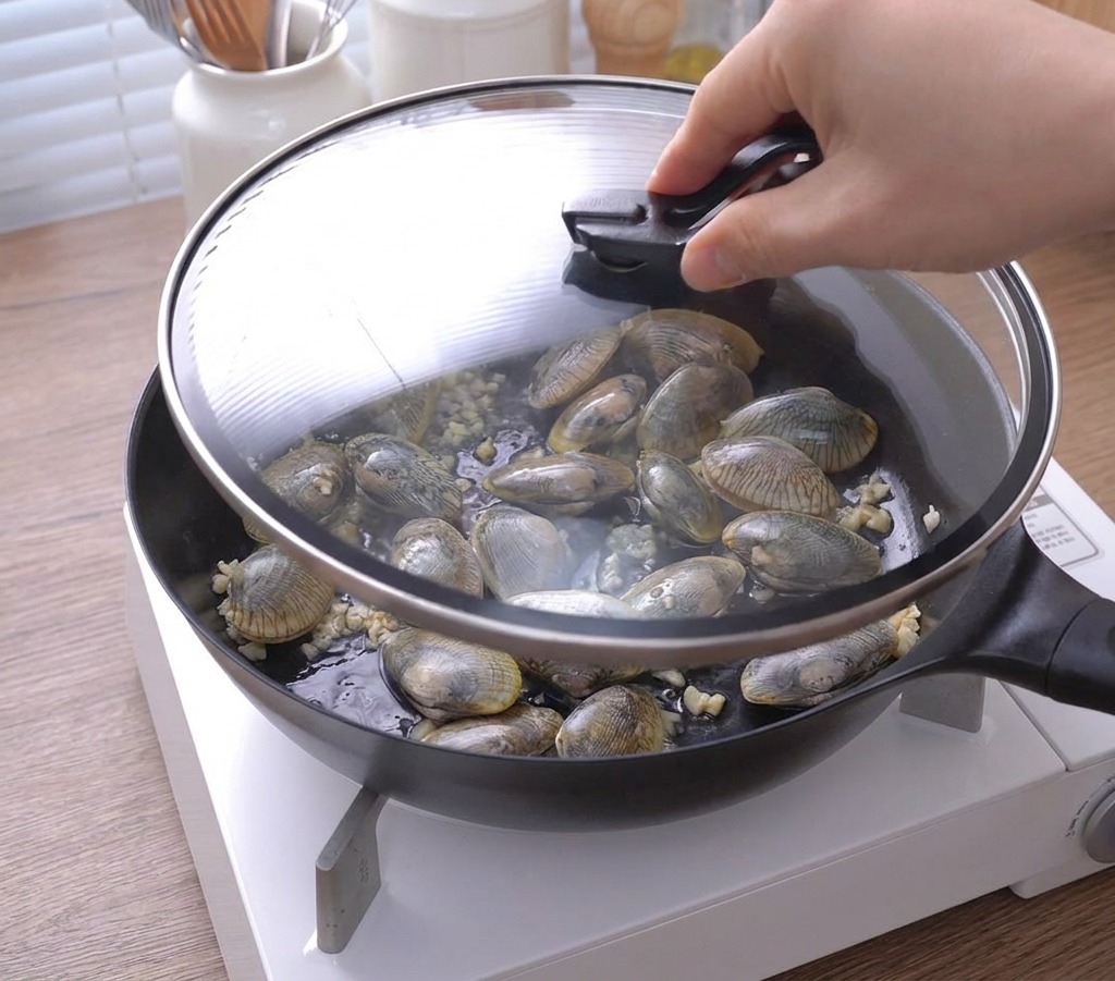 Eine Hand setzt einen Glasdeckel auf die Pfanne mit den Muscheln und dem Knoblauch.