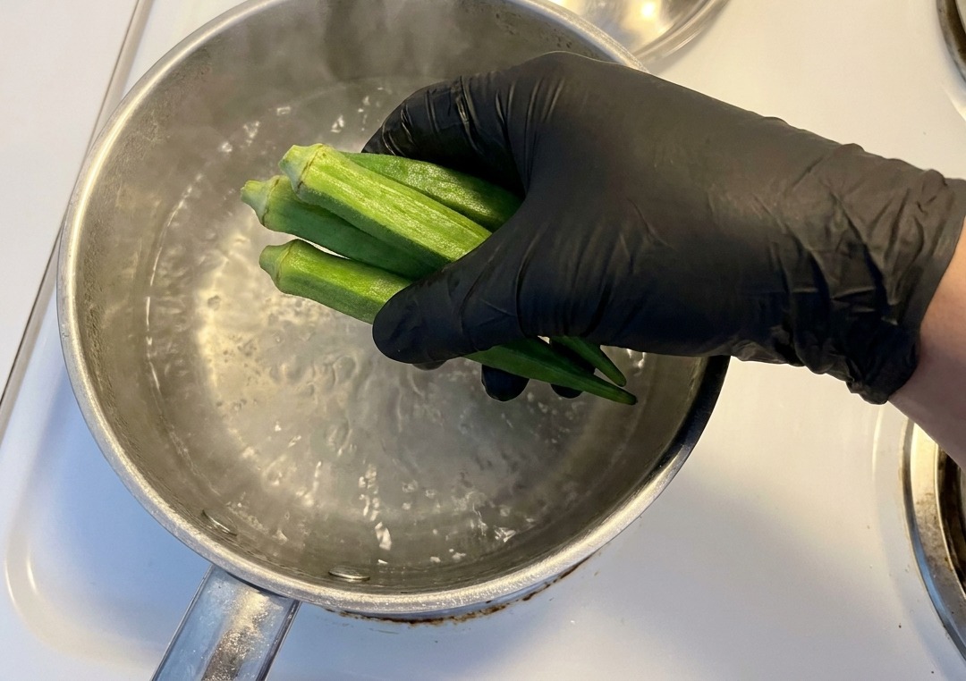 Eine Hand in Handschuhen gibt frische, ganze Okraschoten in einen Edelstahltopf mit kochendem Wasser auf einem weißen Herd.