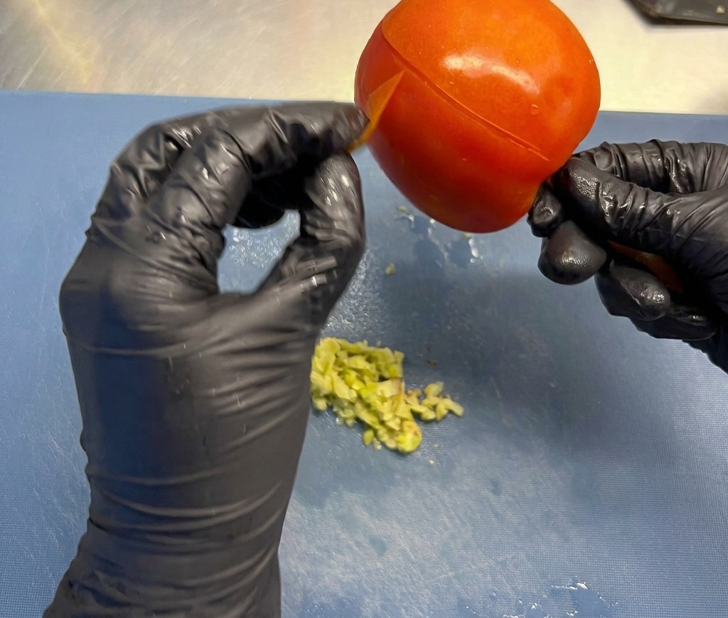 Hände in schwarzen Handschuhen ziehen die dünne Haut einer großen, reifen roten Tomate über einem Brett ab.