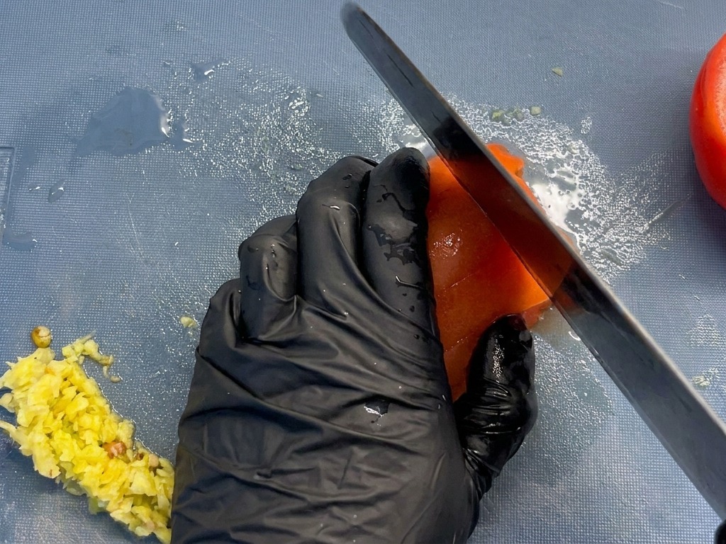 Nahaufnahme eines Kochs, der eine gehäutete rote Tomate in gleichmäßige Stücke auf einem blauen Brett schneidet.