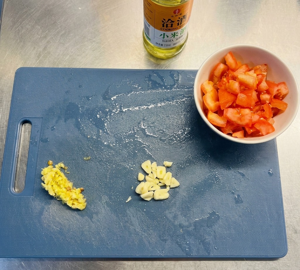 Ein blaues Schneidebrett mit gehacktem Knoblauch, Peperoni und Tomatenwürfeln neben einer Flasche Öl.