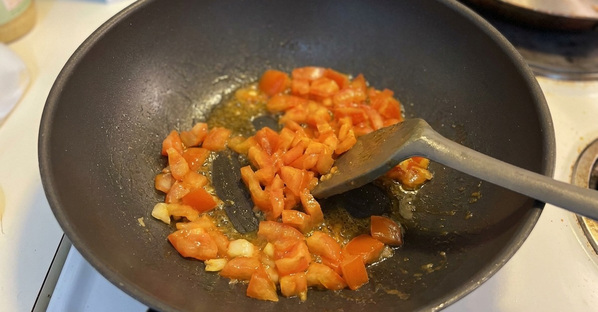 Tomatenwürfel und Knoblauch werden in einem dunklen Wok angeschwitzt, während der Saft am Boden zu blubbern beginnt.