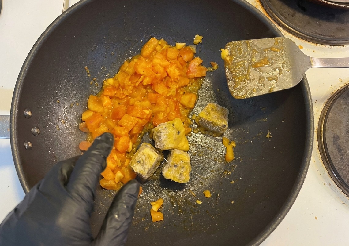 Gefrorene Passionsfrucht-Würfel und gehackte Peperoni werden von einer Hand in die Pfanne zu den Tomaten gegeben.