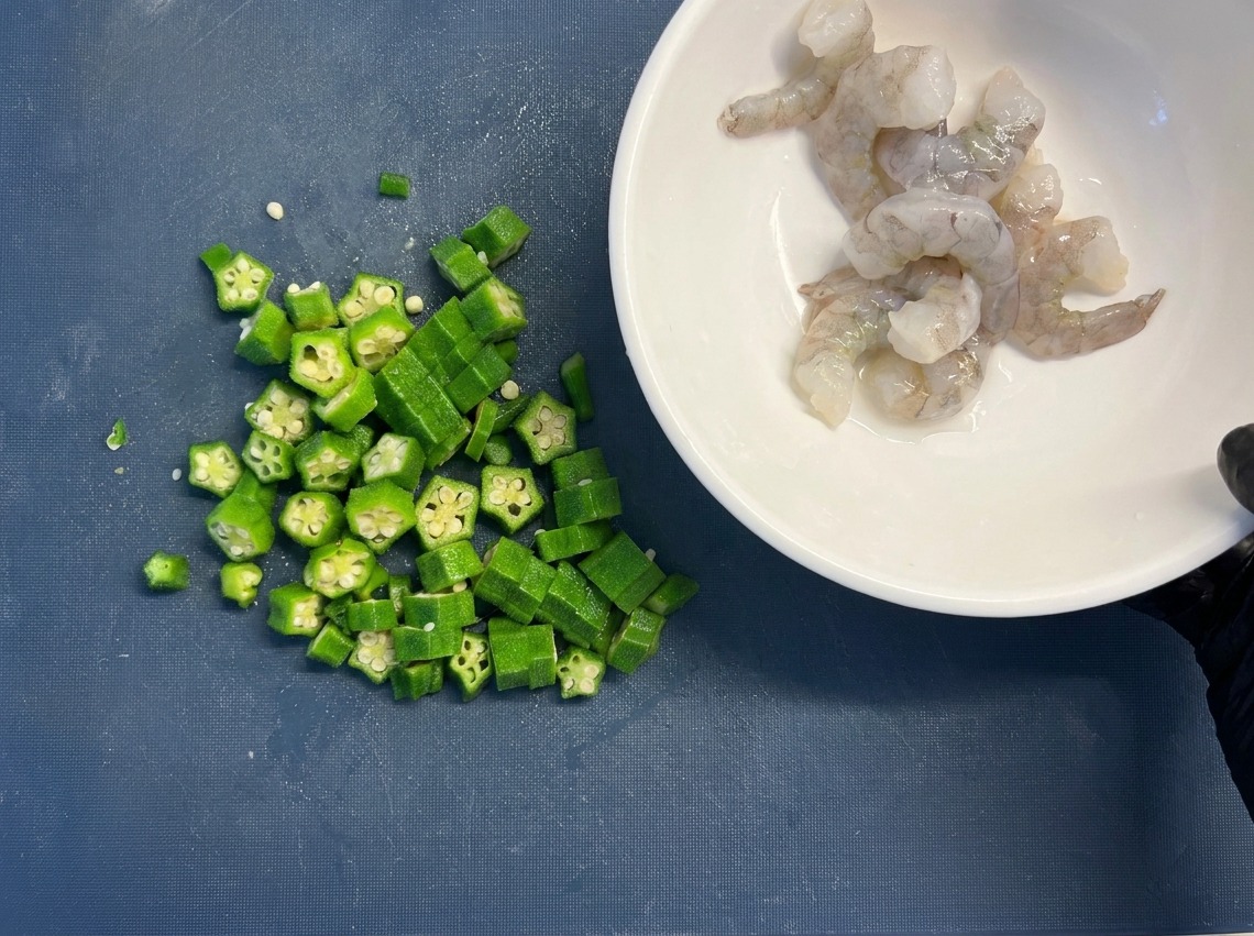 Eine weiße Schüssel mit rohen Garnelen steht neben einem Haufen grüner Okra-Ringe auf einem Schneidebrett.