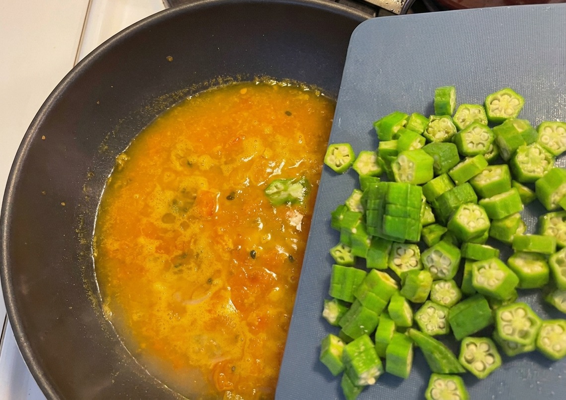 Frische Okra-Ringe werden von einem blauen Schneidebrett in die köchelnde orangefarbene Suppe im Wok gegeben.