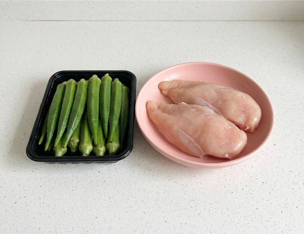 Ein schwarzes Tablett mit frischen Okraschoten neben zwei rohen Hähnchenbrustfilets auf einer weißen Arbeitsplatte.