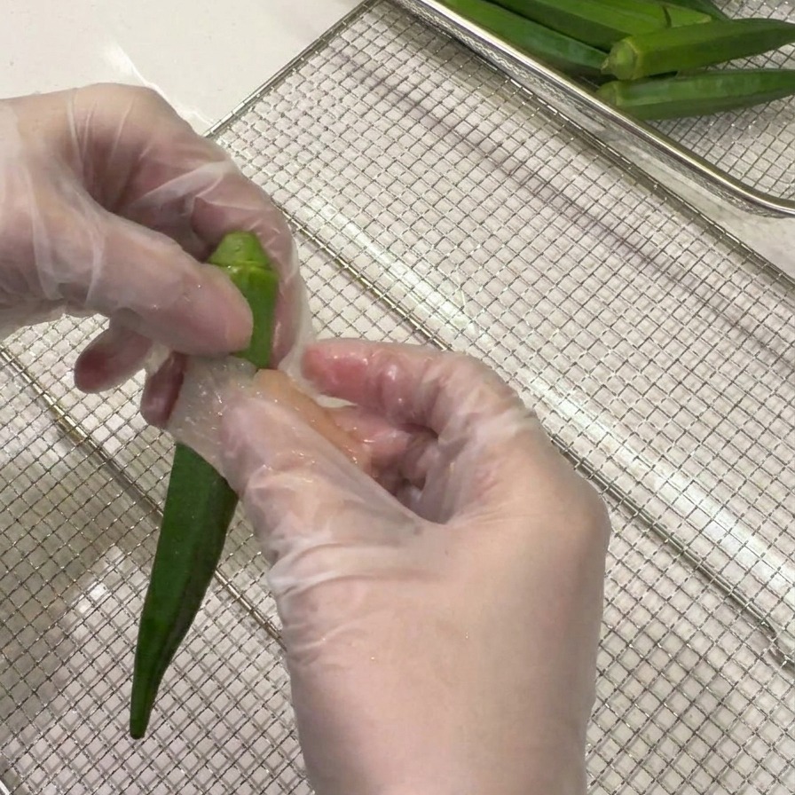 Hände mit Handschuhen umwickeln eine dünne Scheibe rohe Hähnchenbrust spiralförmig um eine ganze grüne Okraschote.