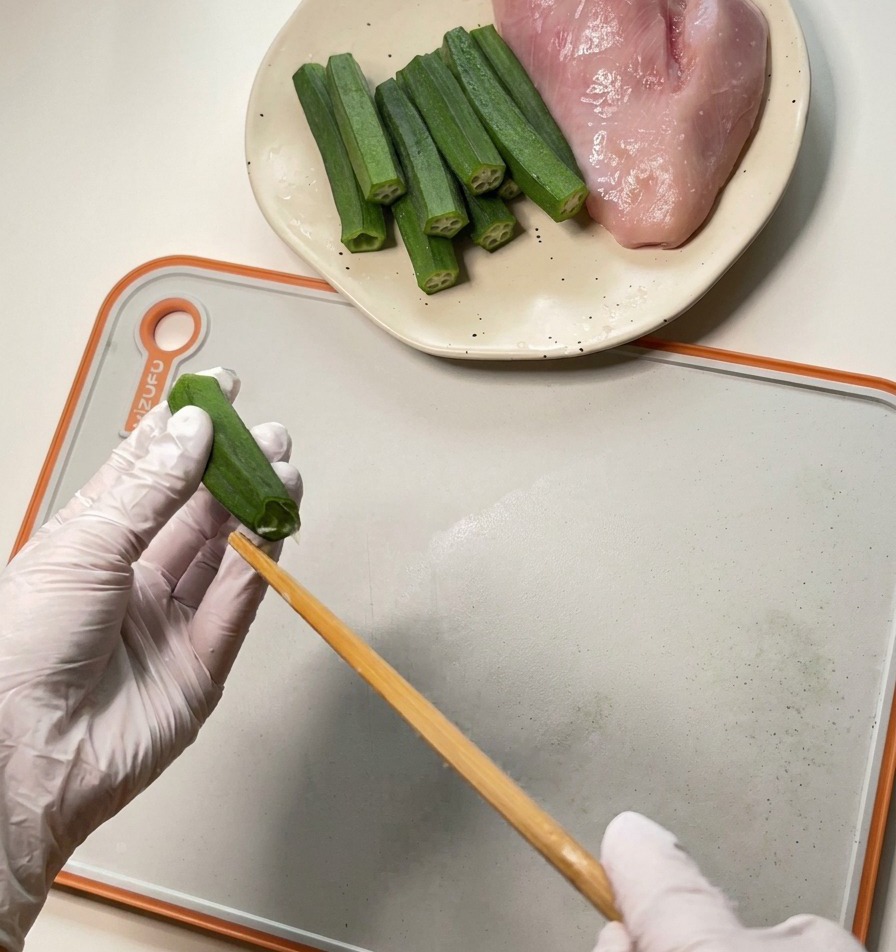 Eine Person mit weißen Handschuhen drückt mit einem Holzstäbchen Kerne und Fruchtfleisch aus einer grünen Okraschote.