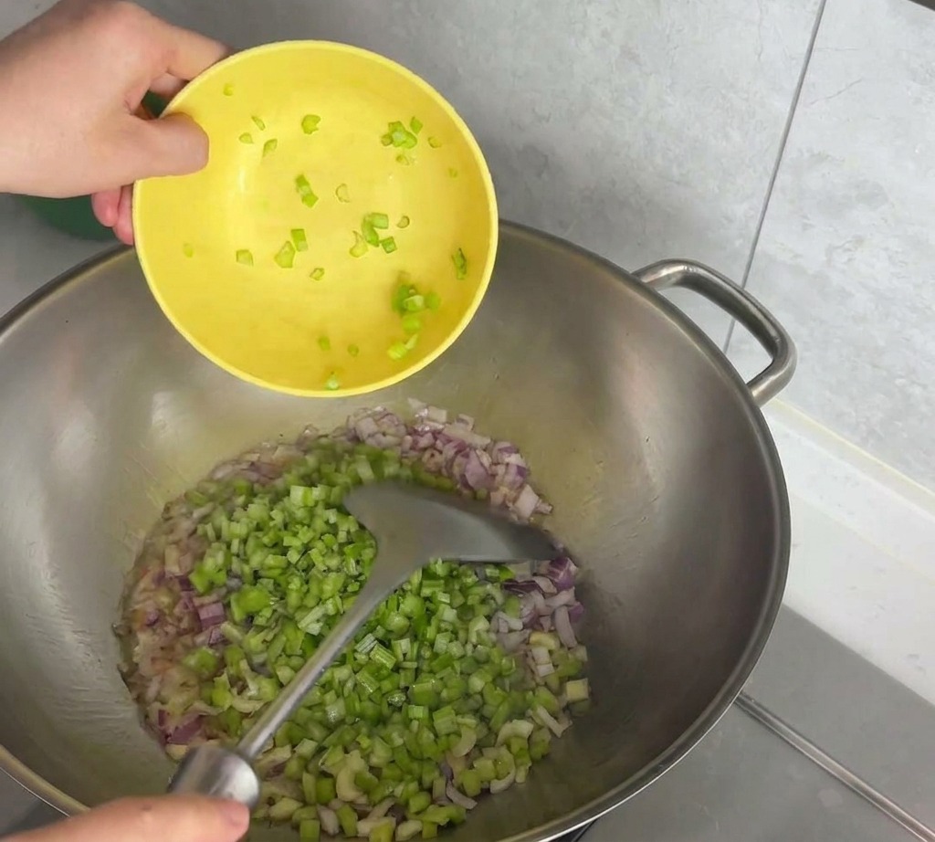 Eine Person gibt gewürfelten Sellerie aus einer gelben Schüssel in eine Edelstahlpfanne mit gehackten roten Zwiebeln.