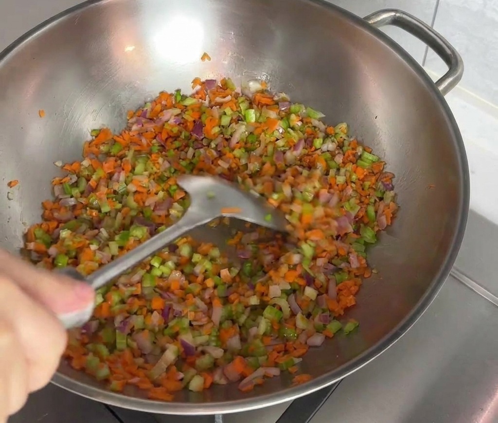 Eine bunte Mischung aus fein gewürfelten Karotten, Sellerie und roten Zwiebeln wird in einem Edelstahl-Wok mit einem Pfannenwender angebraten.