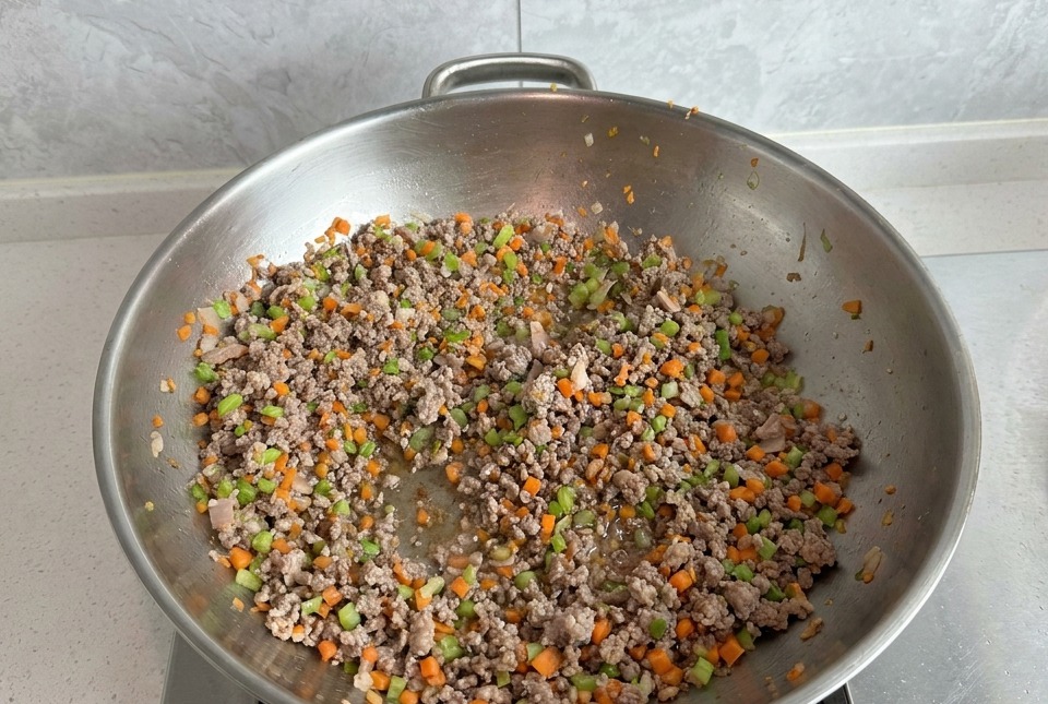 Hackfleisch wird mit gewürfelten Karotten und Sellerie in einem großen silbernen Wok angebraten; das Fleisch ist bereits deutlich gebräunt.