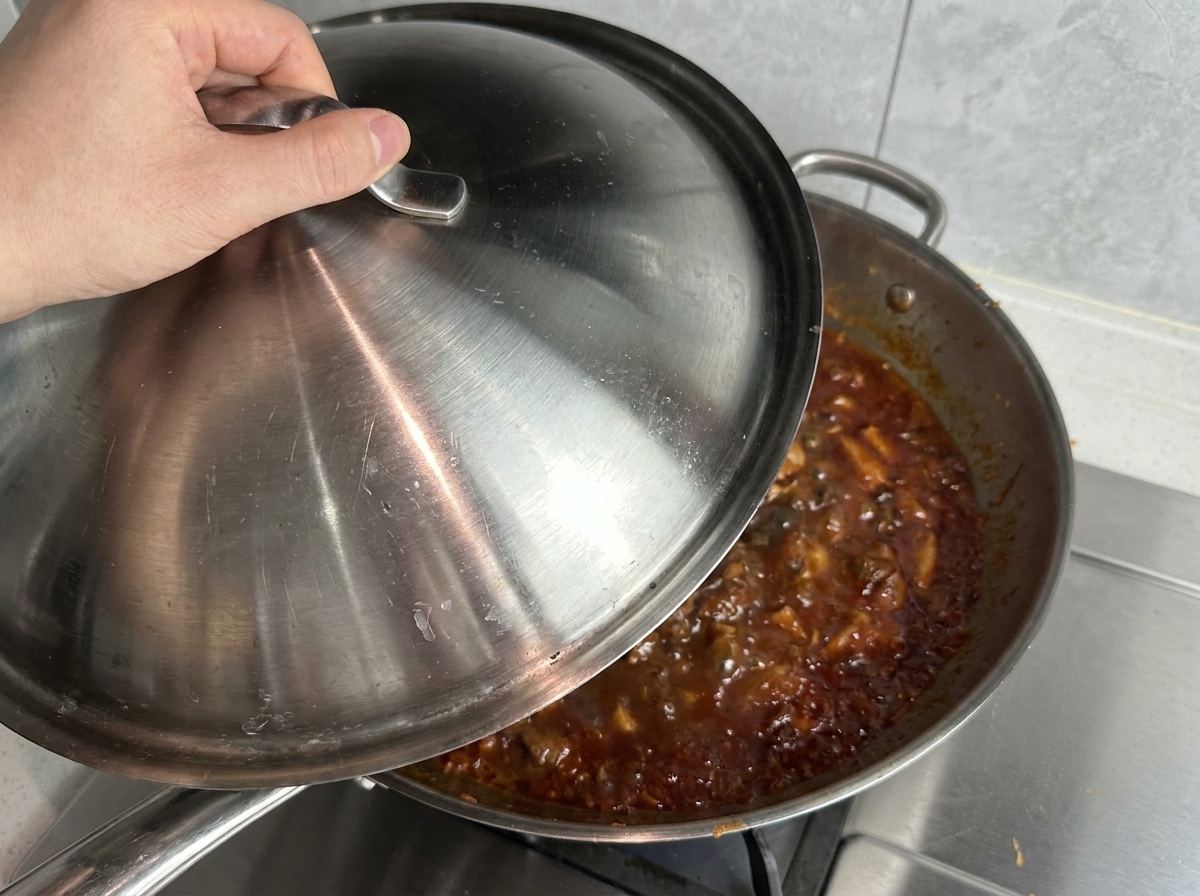 Eine Hand legt einen Edelstahldeckel auf eine große Wok-Pfanne, in der die rote Fleischsauce blubbert.