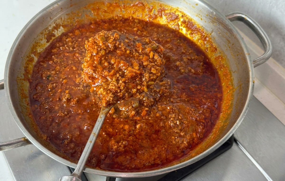 Eine Edelstahlpfanne mit dicker, dunkelroter Bolognese-Sauce; ein großer Metalllöffel zeigt die sämige Konsistenz.