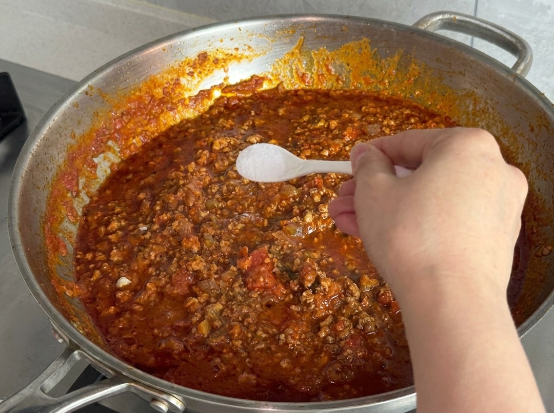 Eine Hand hält einen kleinen weißen Plastiklöffel mit feinem Salz über die köchelnde Sauce.