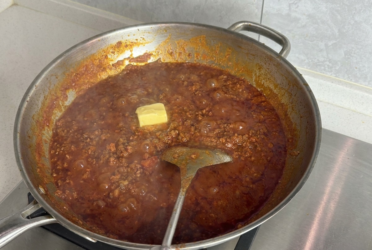Ein quadratisches Stück Butter schmilzt auf der Oberfläche einer köchelnden roten Fleischsauce in einer silbernen Pfanne.
