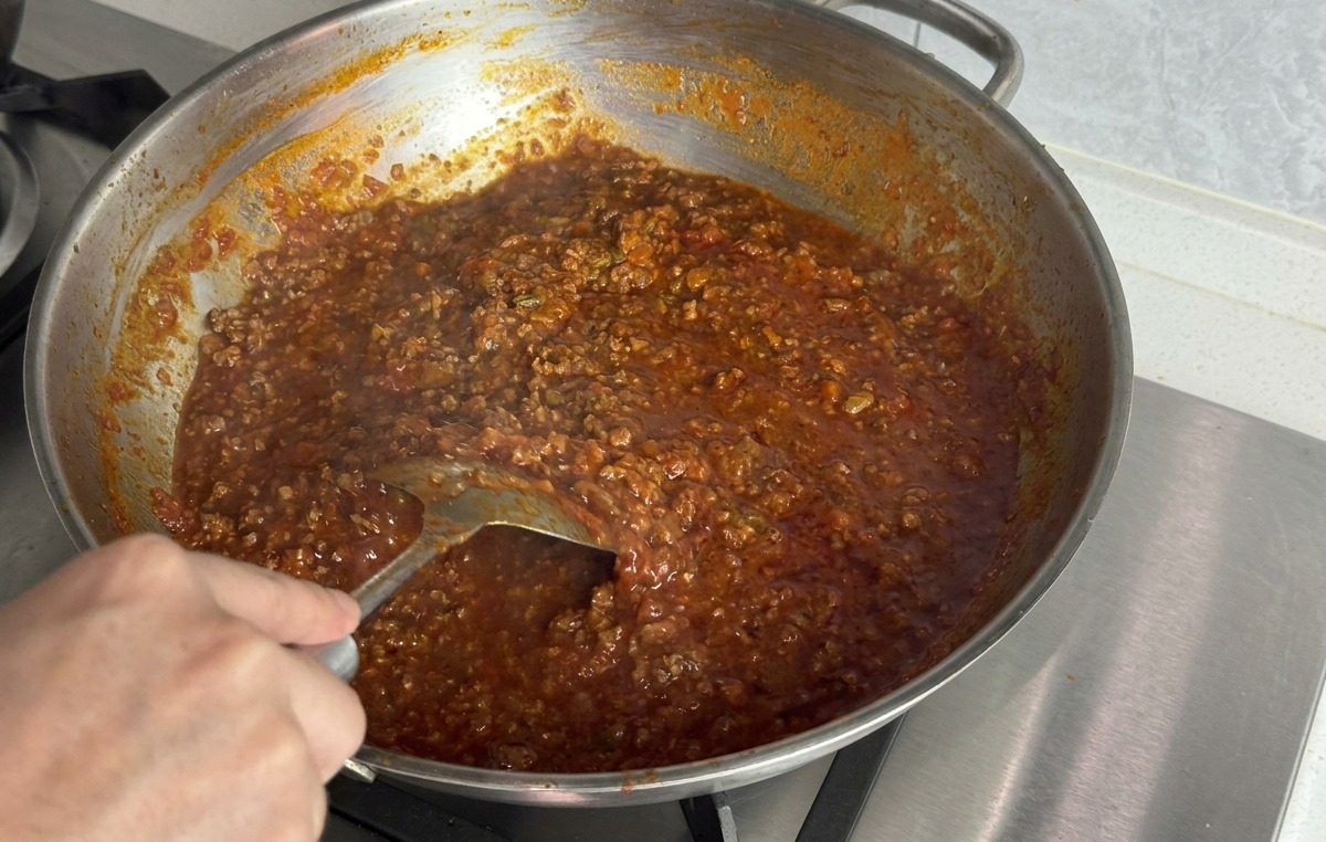 Eine Hand rührt mit einem Metallspatel ein schmelzendes Stück Butter in die dicke rote Fleischsauce ein.
