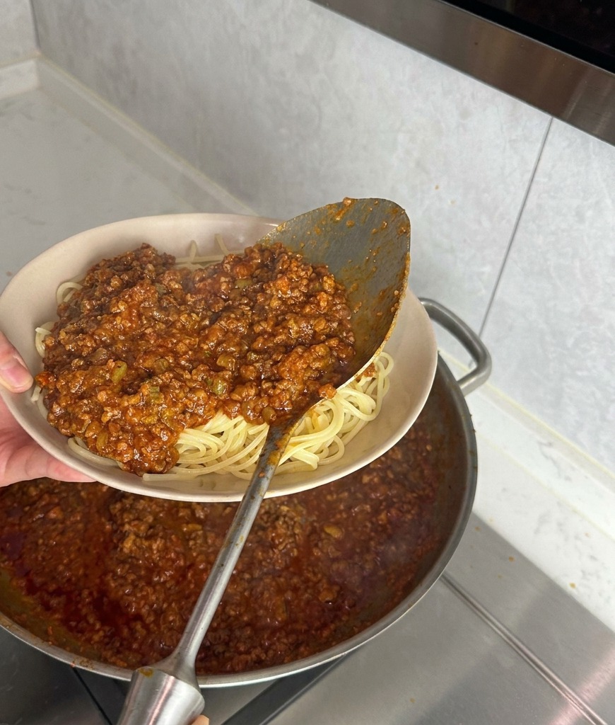 Eine große Metallkelle gießt dicke Fleischsauce über eine Portion schlicht gekochte Spaghetti in einer beigen Schüssel.