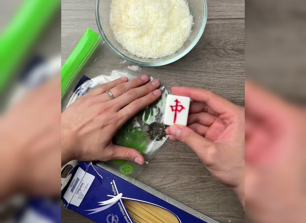 Eine Hand, die mit der flachen Seite eines Mahjong-Steins schwarze Pfefferkörner in einem Plastikbeutel auf einem Holztisch zerdrückt.