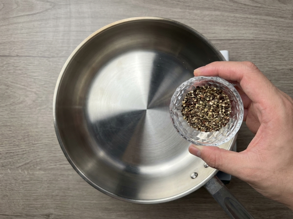 Grob zerstoßener schwarzer Pfeffer wird aus einer kleinen Glasschale in eine trockene, vorgeheizte Edelstahlpfanne geschüttet.