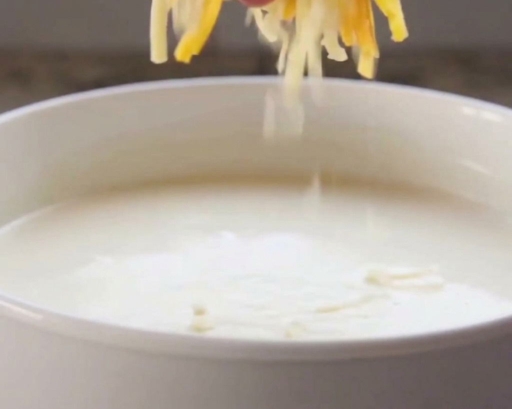Eine Hand gibt geriebenen orangefarbenen und weißen Käse in eine Schüssel mit warmer heller Rahmsauce