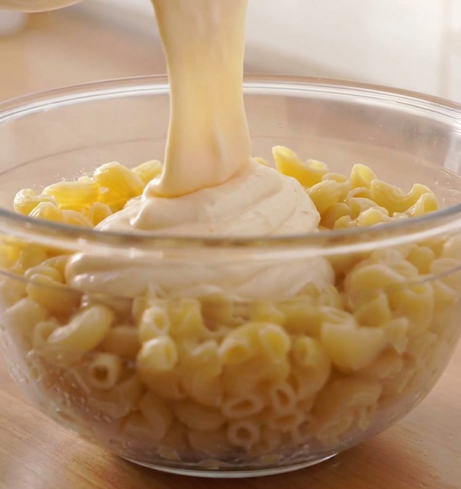 Ein dicker Strahl geschmolzener weißer Käsesauce wird über eine große Schüssel mit gekochten Spiralnudeln gegossen