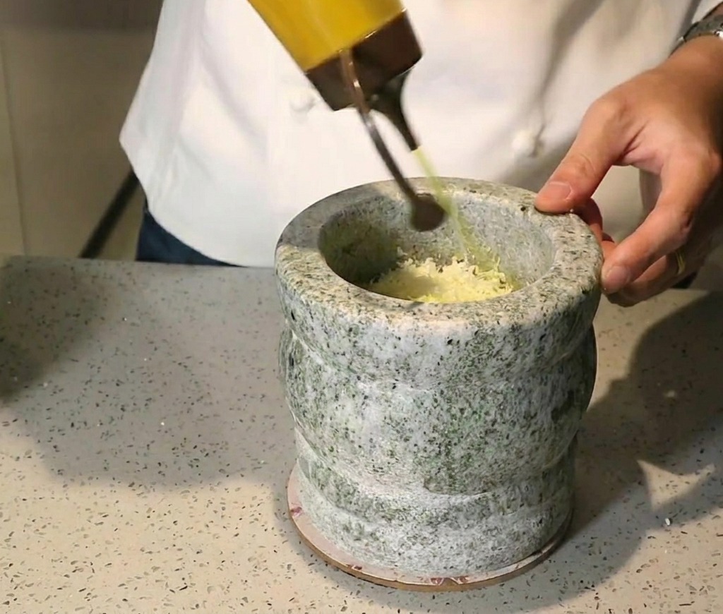 Goldenes Olivenöl wird aus einer Spritzflasche in einen Steinmörser gegossen, der mit zerstoßenen grünen Kräutern und Nüssen gefüllt ist.