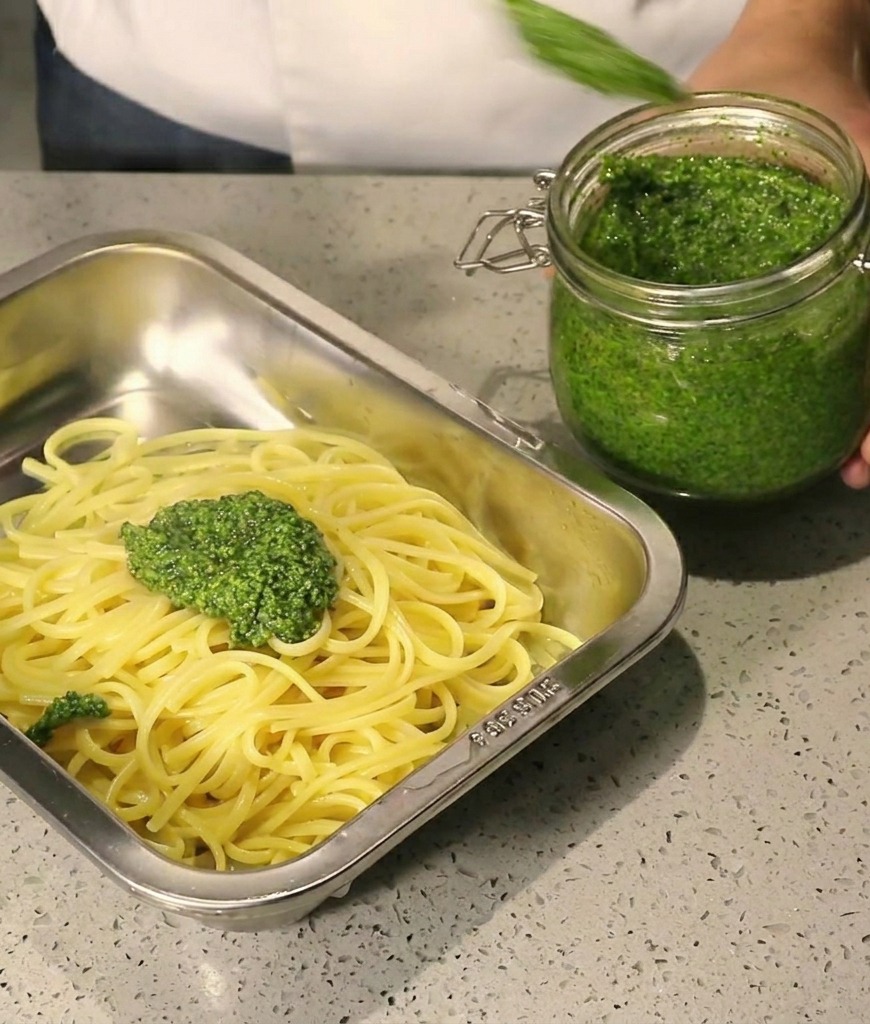 Dickes grünes Pesto wird aus einem Glas auf heiße Spaghetti in einem Edelstahltablett gelöffelt.