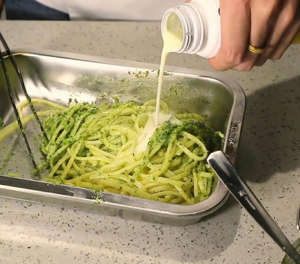 Ein Strahl cremiger weißer Sauce wird auf Pasta gegossen, die mit grünem Pesto bedeckt ist.
