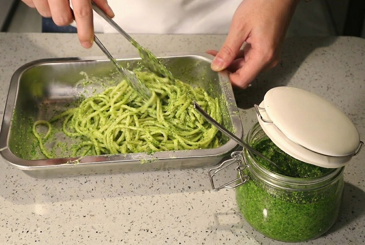 Mit einer Metallzange werden Spaghetti mit grünem Pesto und weißer Sahne in einem Metalltablett verrührt und vermischt.