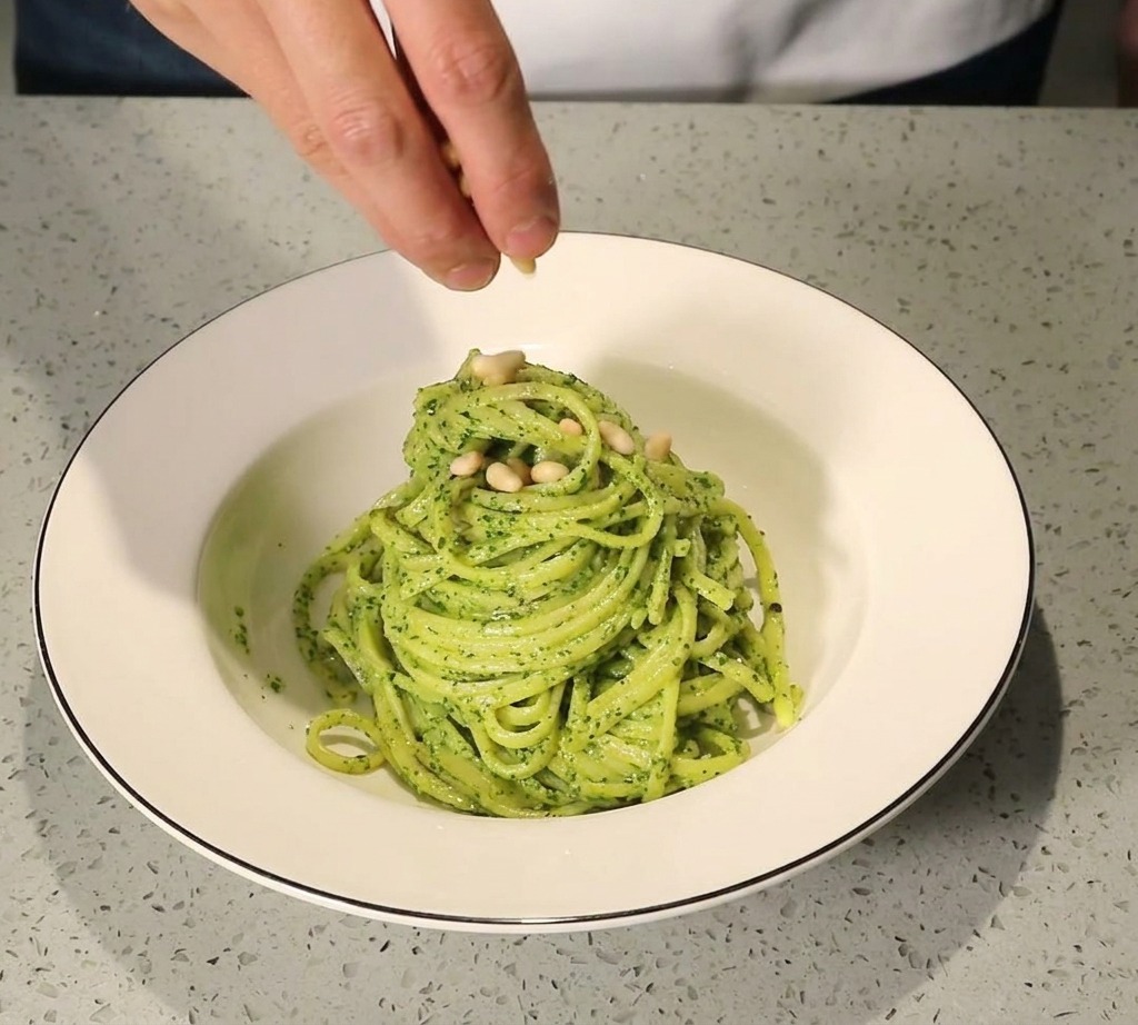 Eine Hand streut goldbraun geröstete Pinienkerne auf eine hübsch angerichtete Portion Pesto-Pasta in einer weißen Schüssel.