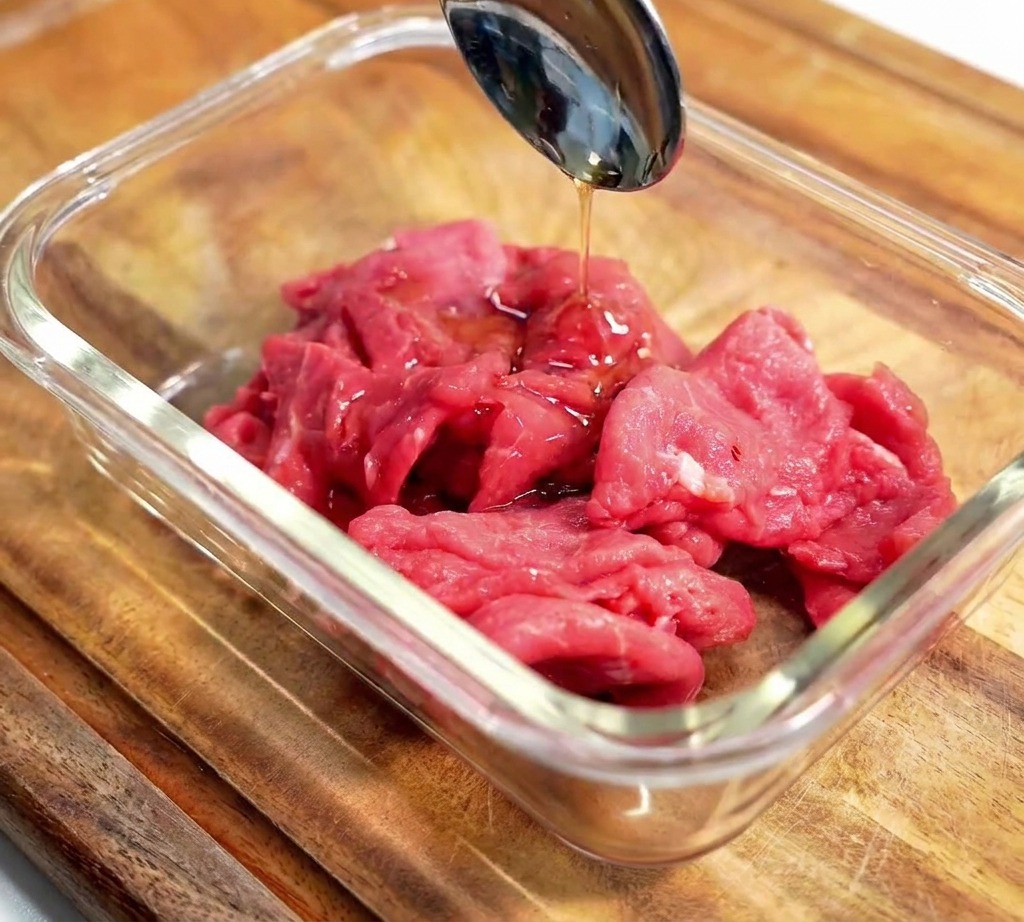 Ein Metalllöffel gießt dunkle Marinade über rohe Rindfleischscheiben in einer Glasschüssel.