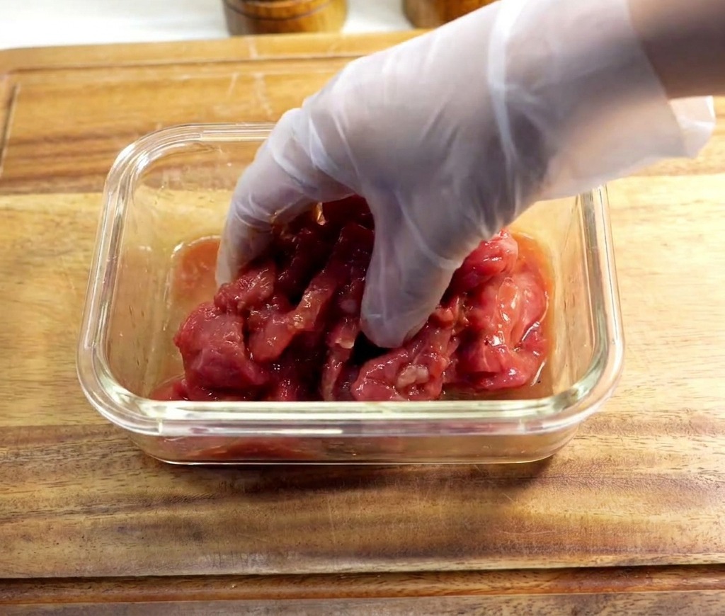 Eine Hand mit durchsichtigem Handschuh knetet die Marinade in die rohen Fleischscheiben ein.