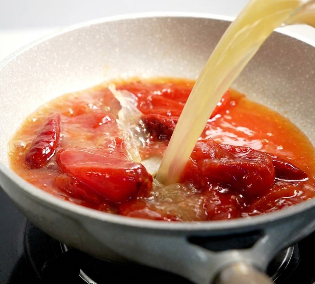 Brühe wird in eine Pfanne mit einer blubbernden, roten Tomatensaucen-Mischung gegossen.