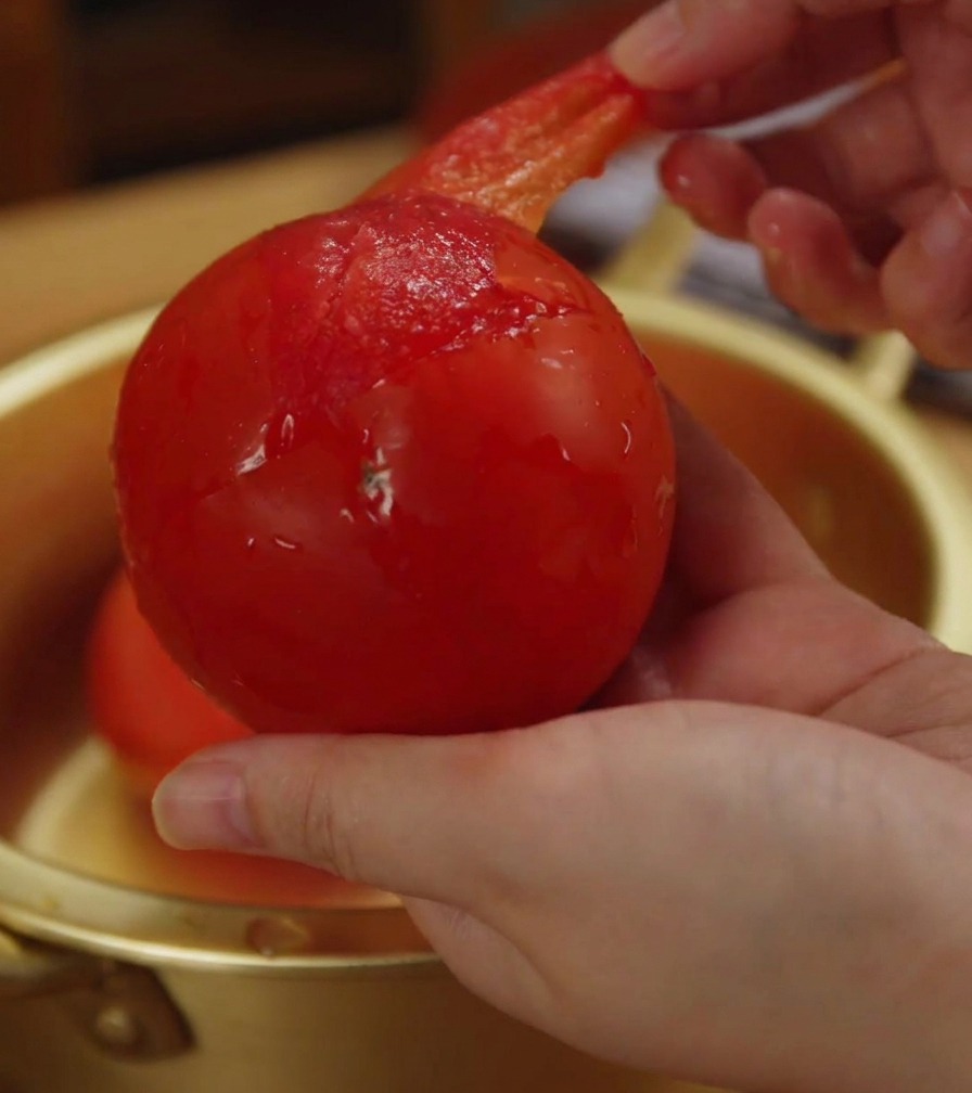 Nahaufnahme einer Hand, die die dünne, durchscheinende rote Schale von einer weichen Tomate abzieht.