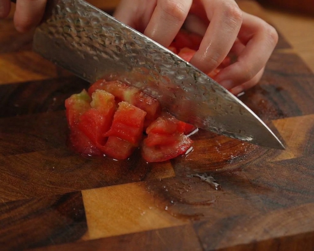 Eine Person würfelt eine geschälte rote Tomate auf einem dunklen Holzbrett mit einem Küchenmesser.