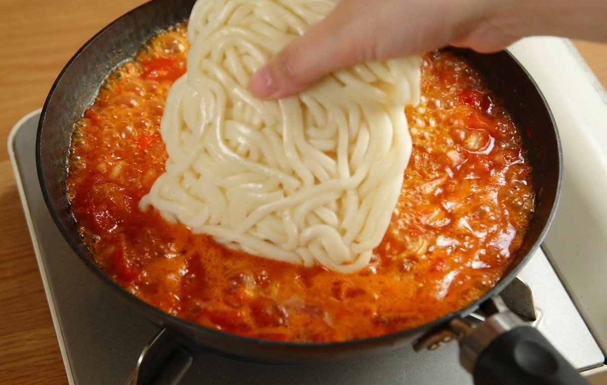 Eine Hand legt einen quadratischen Block weißer Udon-Nudeln in eine Pfanne mit köchelnder Tomatensauce.
