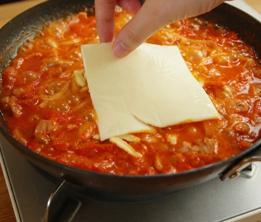 Eine Hand legt eine quadratische Scheibe gelben Schmelzkäse auf schmelzenden Mozzarella und Udon-Nudeln.