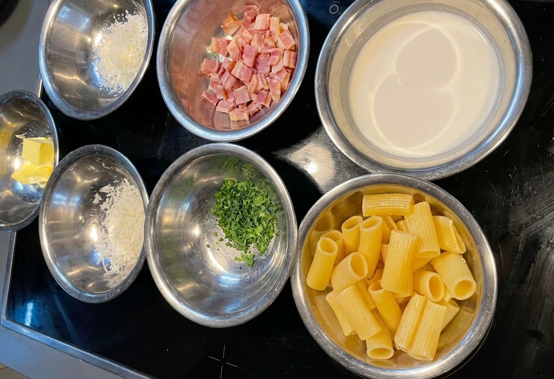 Verschiedene Edelstahlschalen mit abgemessenen Portionen Rigatoni, Sahne, Speck, Käse und Petersilie auf einem dunklen Herd.