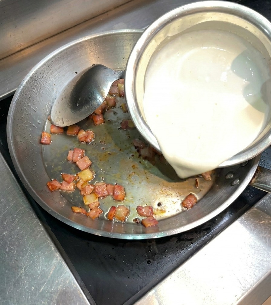 Weiße Sahne wird in eine Edelstahlpfanne zu dem knusprig gebratenen Speck gegossen.