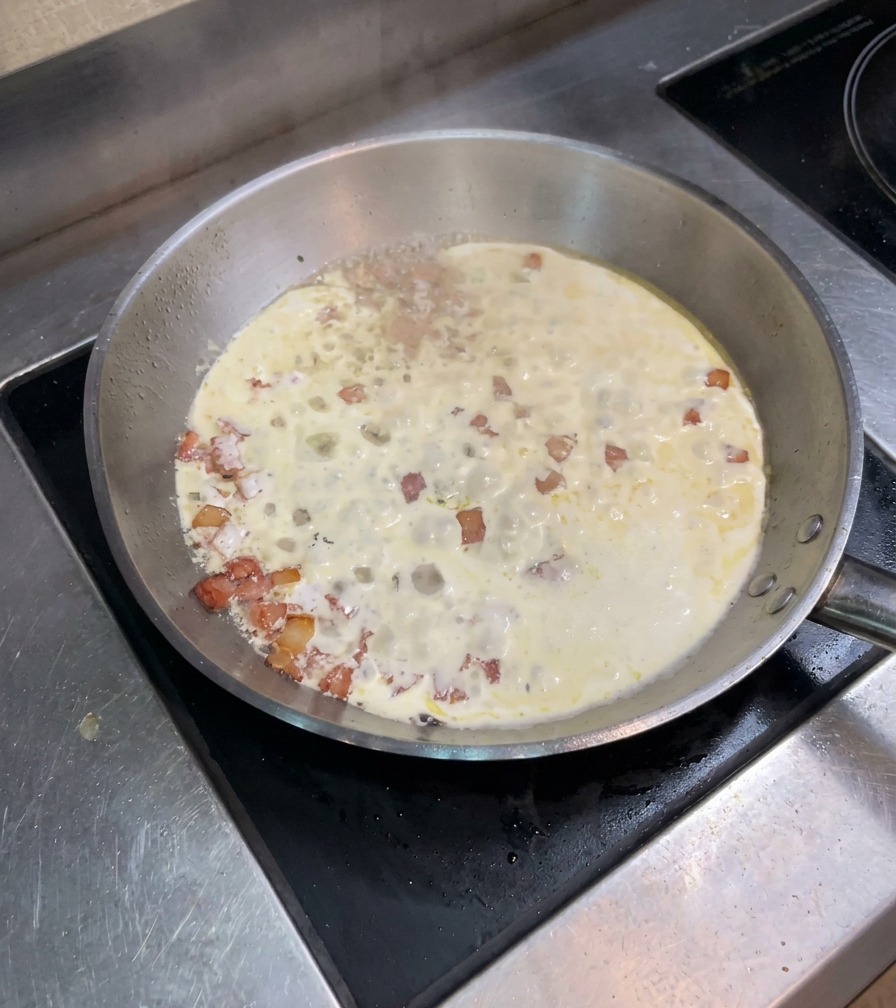Eine Mischung aus Sahne und Speck köchelt sprudelnd in einer breiten Pfanne auf dem Herd.