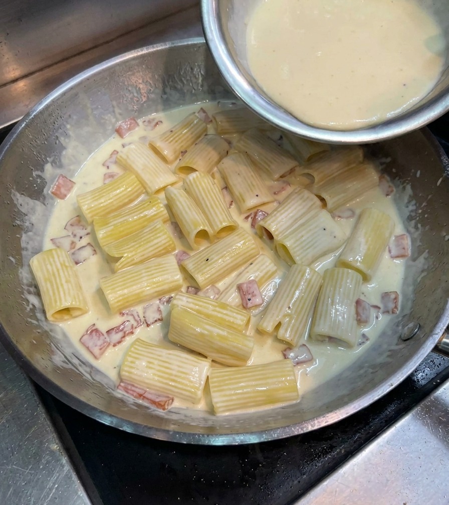 Eine gelbe Eigelb-Käse-Mischung wird über die Rigatoni in der großen Metallpfanne gegossen.