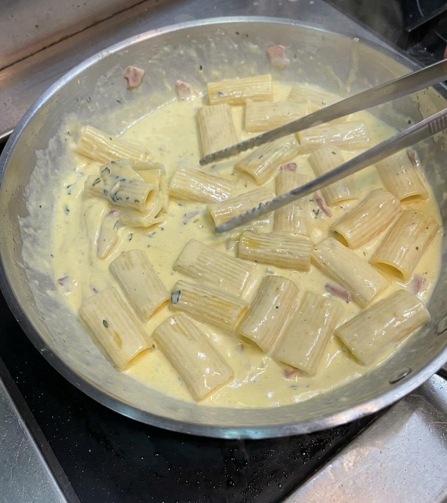 Eine Zange wendet die Rigatoni in der Pfanne, um sie gleichmäßig mit der dicken Carbonara-Soße zu überziehen.