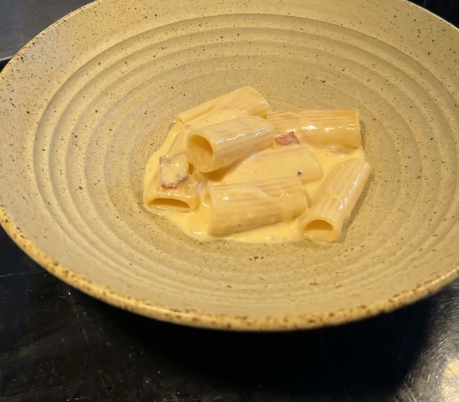 Eine Portion Rigatoni in glänzend gelber Soße mit Speckstücken in einer weiten Keramikschale.