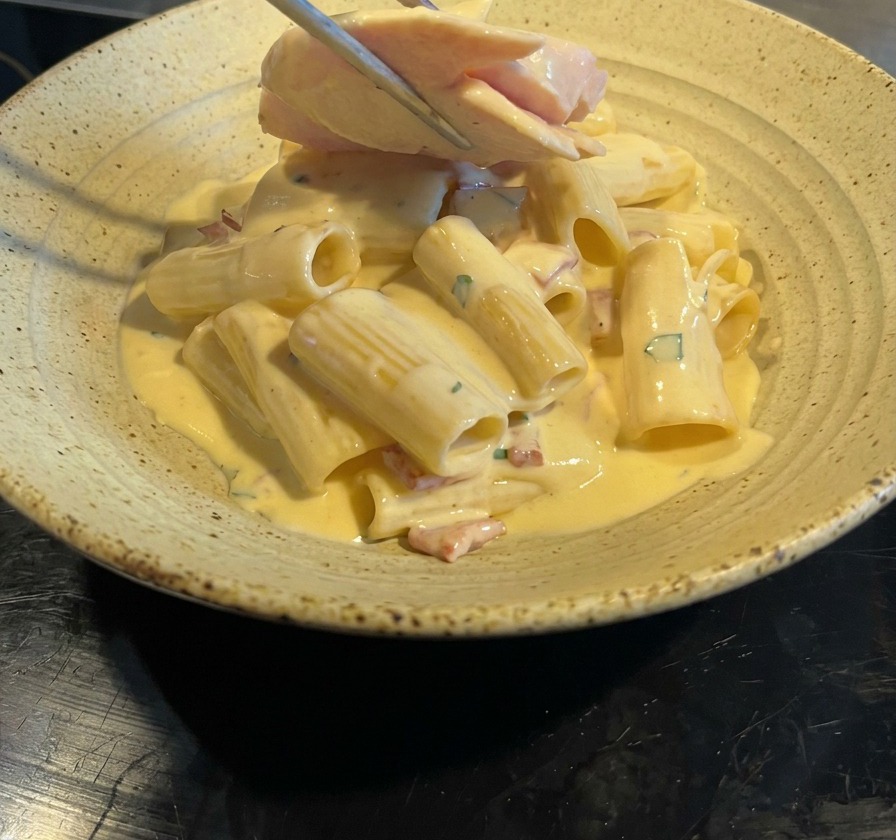 Eine Zange platziert Hähnchenbruststreifen auf den cremigen Rigatoni Carbonara.