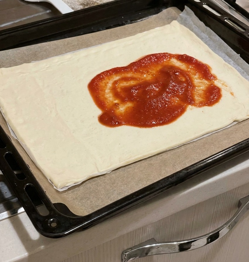 Dickflüssige rote Tomatensauce wird in Klecksen in die Mitte eines rohen rechteckigen Pizzateigs auf einem Blech gegeben.